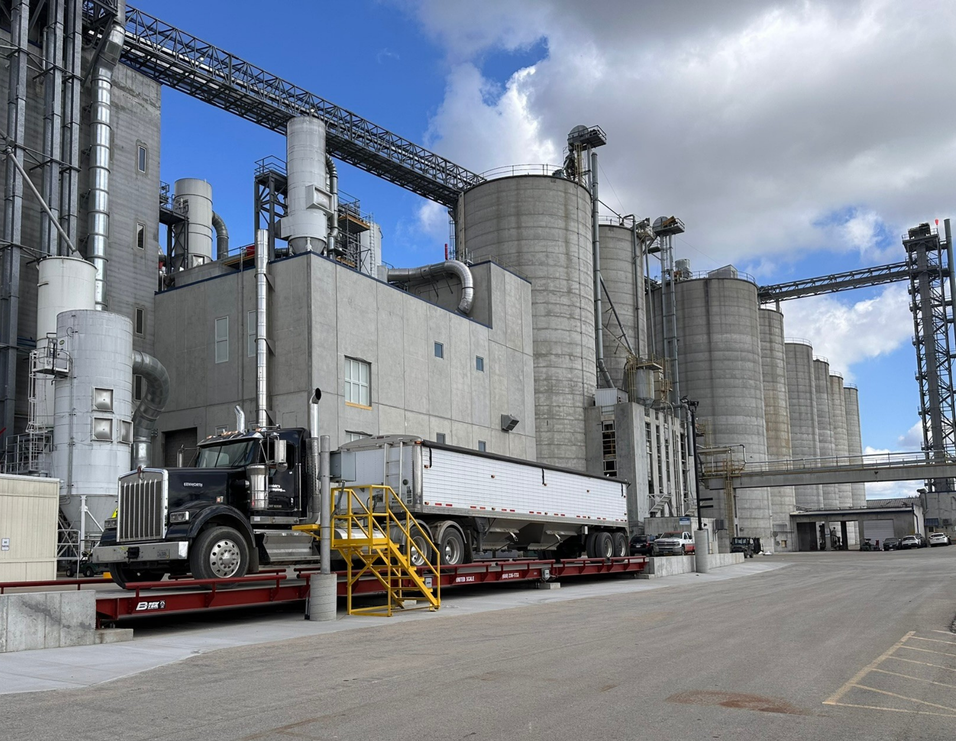 A black semi-truck with a long white trailer is parked on a red industrial weighbridge outside a large concrete grain processing facility, where Masa & Arepa Corn Flour is produced, with silos and pipes in the background under a partly cloudy sky.