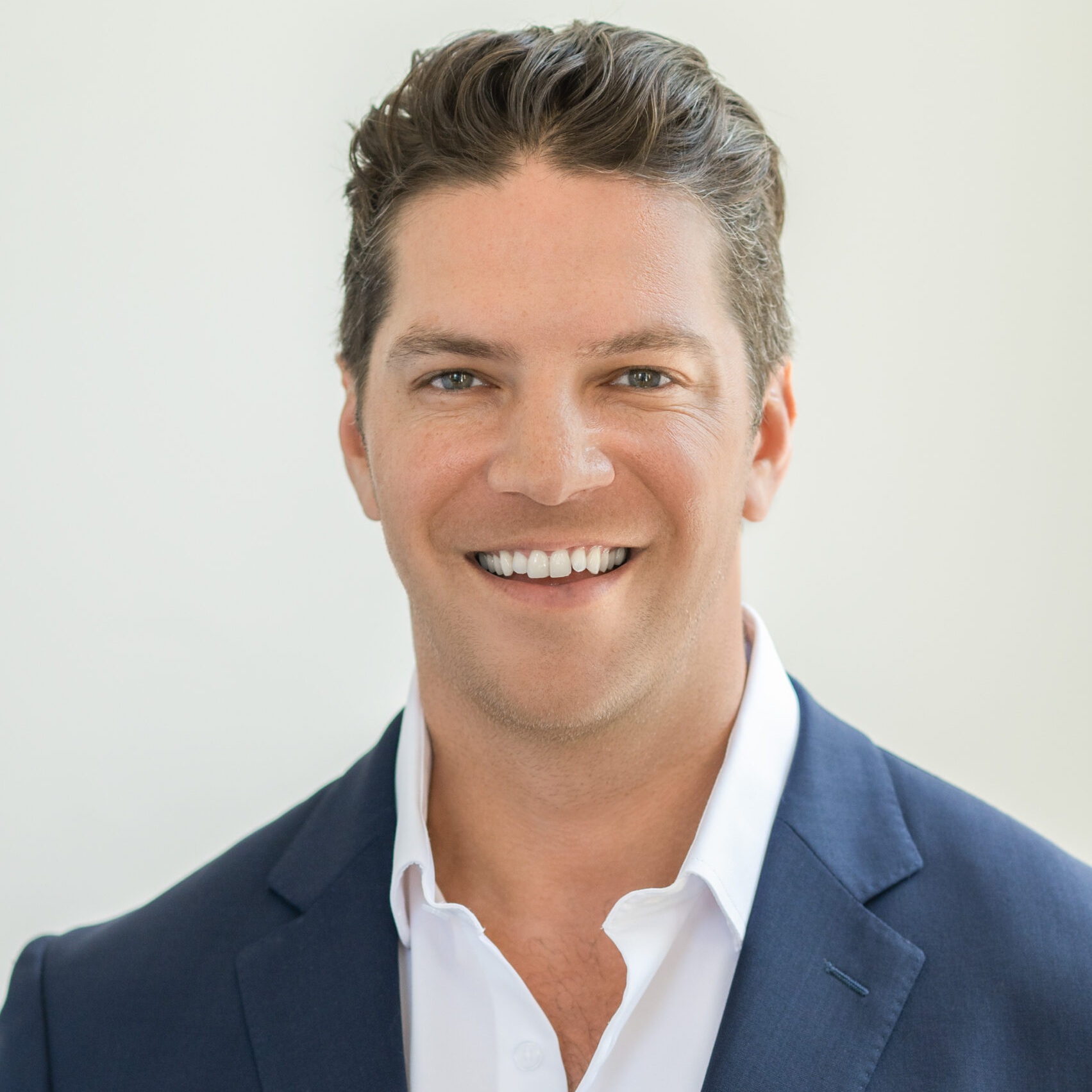 Smiling man with short brown hair wearing a navy blue suit jacket and white dress shirt, standing in front of a plain light background.