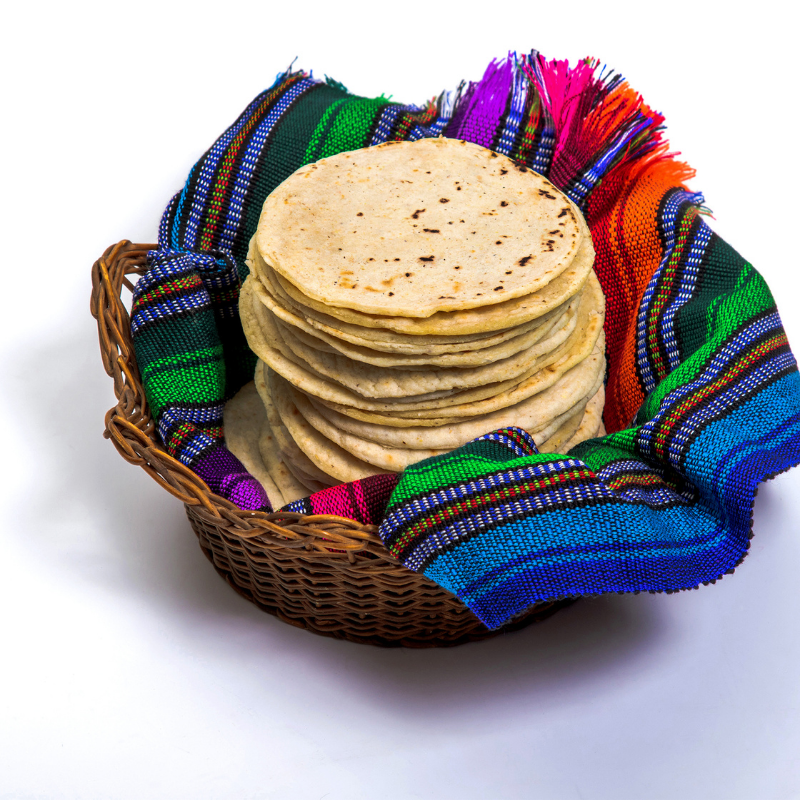 A stack of corn tortillas sits in a woven basket lined with a colorful, traditional Mexican cloth on a white background.
