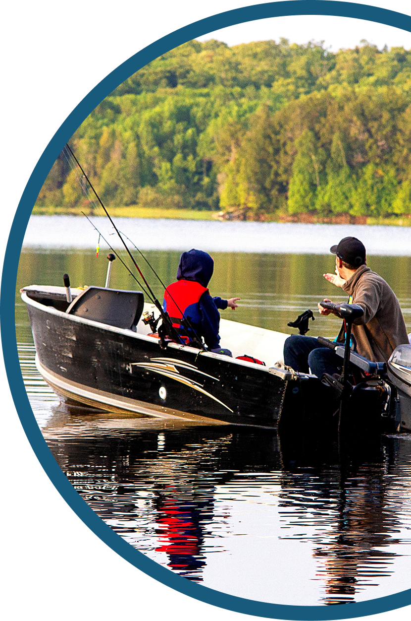 A child in a red and blue jacket and an adult in a cap sit together in a small fishing boat on a calm lake, surrounded by trees, with fishing rods visible.