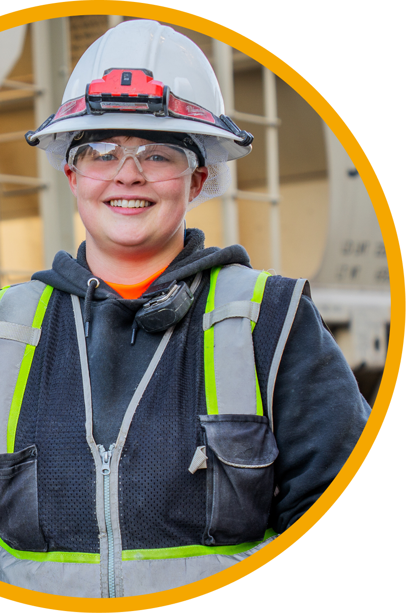 A person wearing a hard hat, safety goggles, and reflective vest smiles at the camera. Industrial equipment is visible in the background.