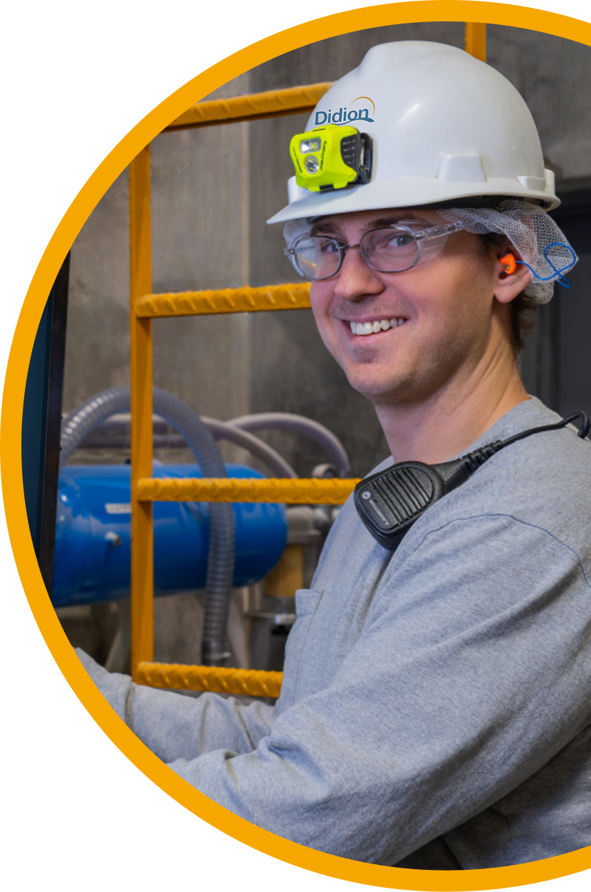 A smiling worker wearing a hard hat with a headlamp, safety glasses, earplugs, and a radio stands in front of industrial equipment and a yellow ladder.