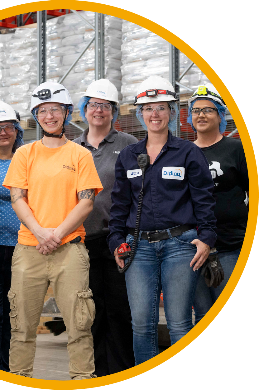 A group of five people wearing hard hats, safety glasses, and work uniforms stand smiling in a warehouse with shelves and stacked bags in the background.