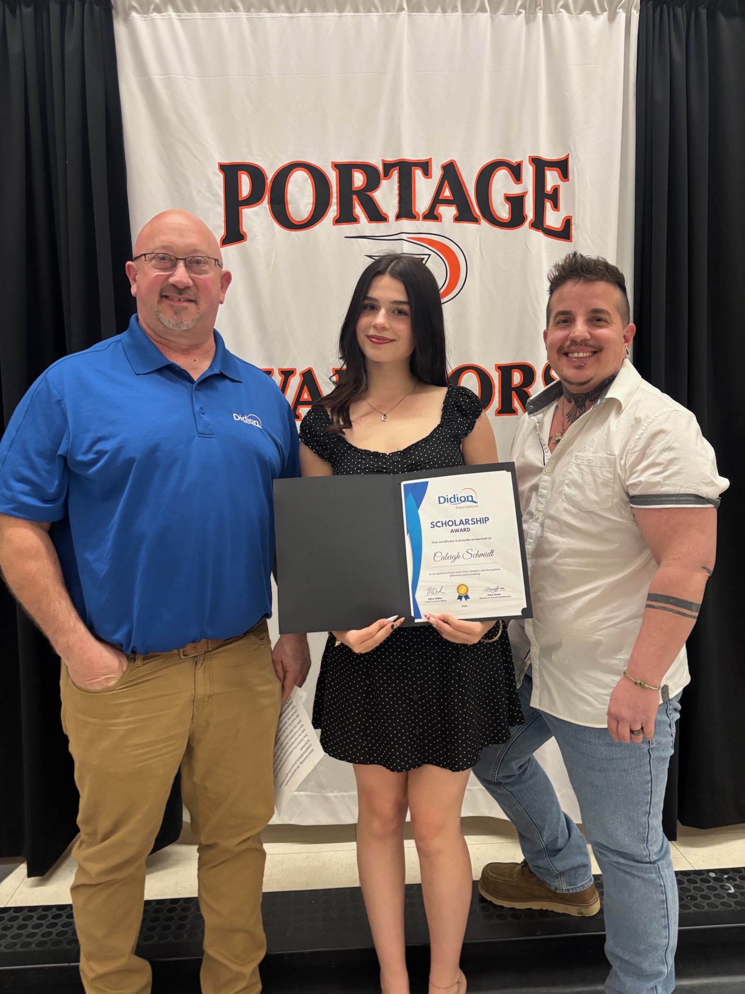 Three people stand in front of a PORTAGE WARRIORS banner. The woman in the center holds a scholarship certificate and a black folder, smiling, with two men, one on each side of her, also smiling at the camera.