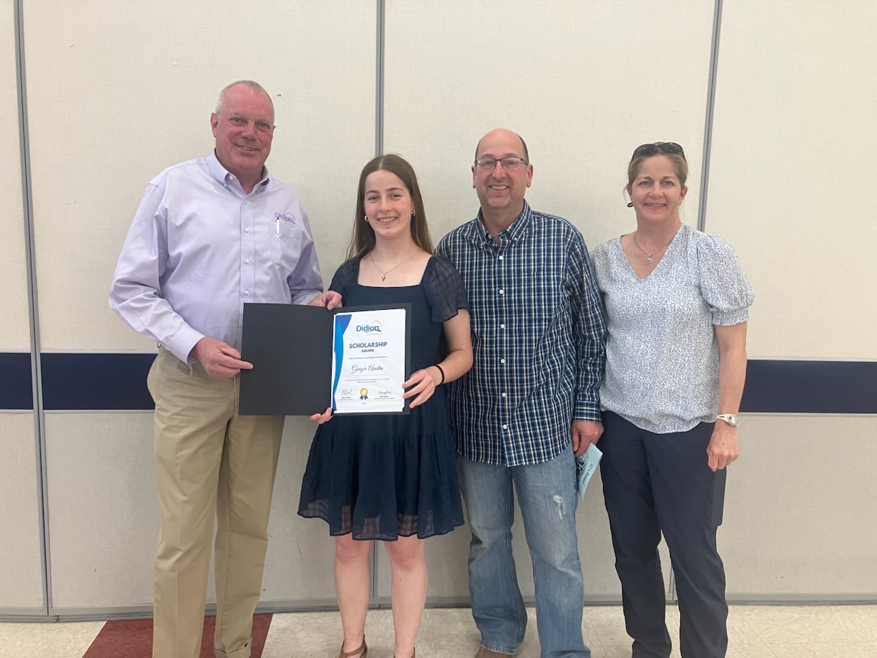 Four people stand indoors, smiling at the camera. A young woman in the center holds a scholarship certificate. Two adults stand on her right, and one adult stands on her left, all appearing happy and supportive.