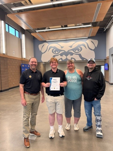 Four people stand indoors, smiling at the camera. The person in the center holds a certificate. Behind them is a large, stylized bulldog mural on the wall. The setting appears to be a school or gymnasium.
