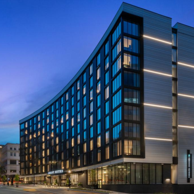 Modern, multi-story hotel with curved glass facade, illuminated exterior lights, and SpringHill Suites sign, photographed at dusk with a blue sky in the background.