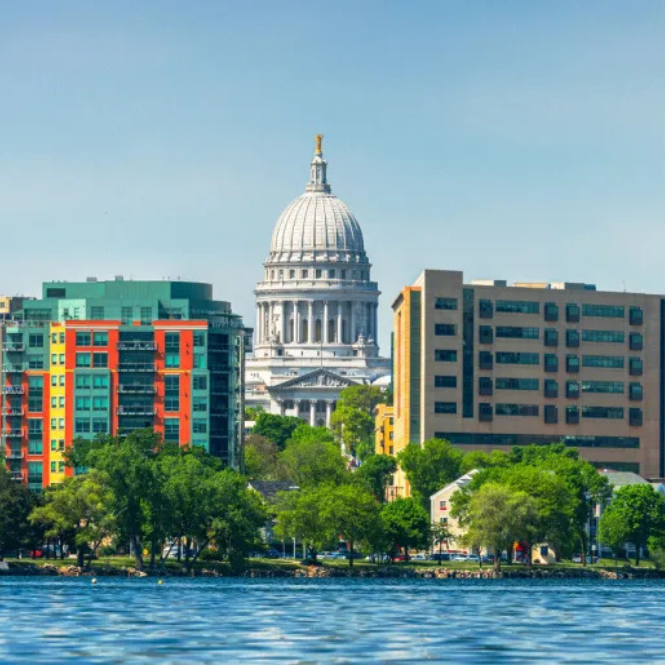 Wisconsin State Capitol building with a white dome, surrounded by colorful modern and older buildings, green trees, and a lake in the foreground on a sunny day.