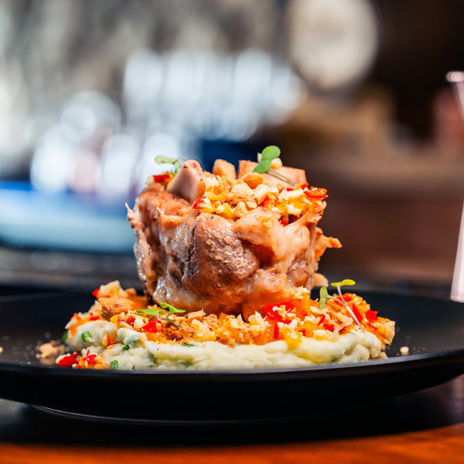 A gourmet dish featuring a stack of tender meat garnished with herbs and crispy bits, served on a bed of mashed potatoes with colorful vegetable pieces, presented on a black plate.