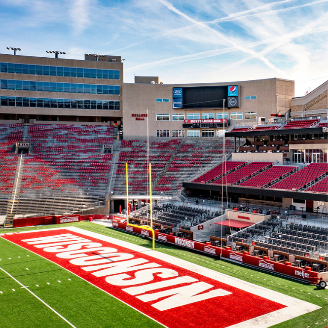 Wisconsin Badgers football stadium with empty red seats, end zone marked WISCONSIN, goalposts, and a scoreboard under a blue sky with streaks of white clouds.