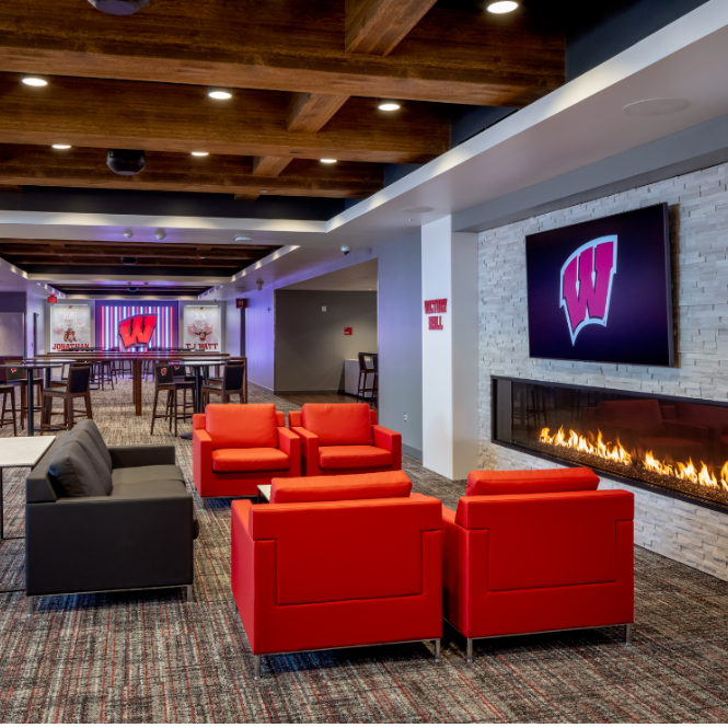 Modern lounge with red and black seating, a fireplace, and large TV screens displaying the Wisconsin Badgers W logo. Exposed wood beams and tall tables are in the background.